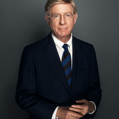White man with blonde hair and glasses in a black suit and tie.